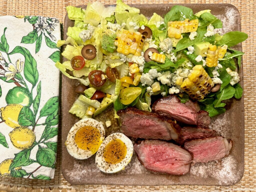 Steak, salad, corn, and eggs on a plate.