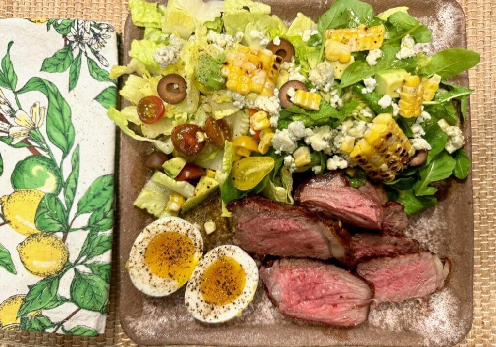 Steak, salad, corn, and eggs on a plate.